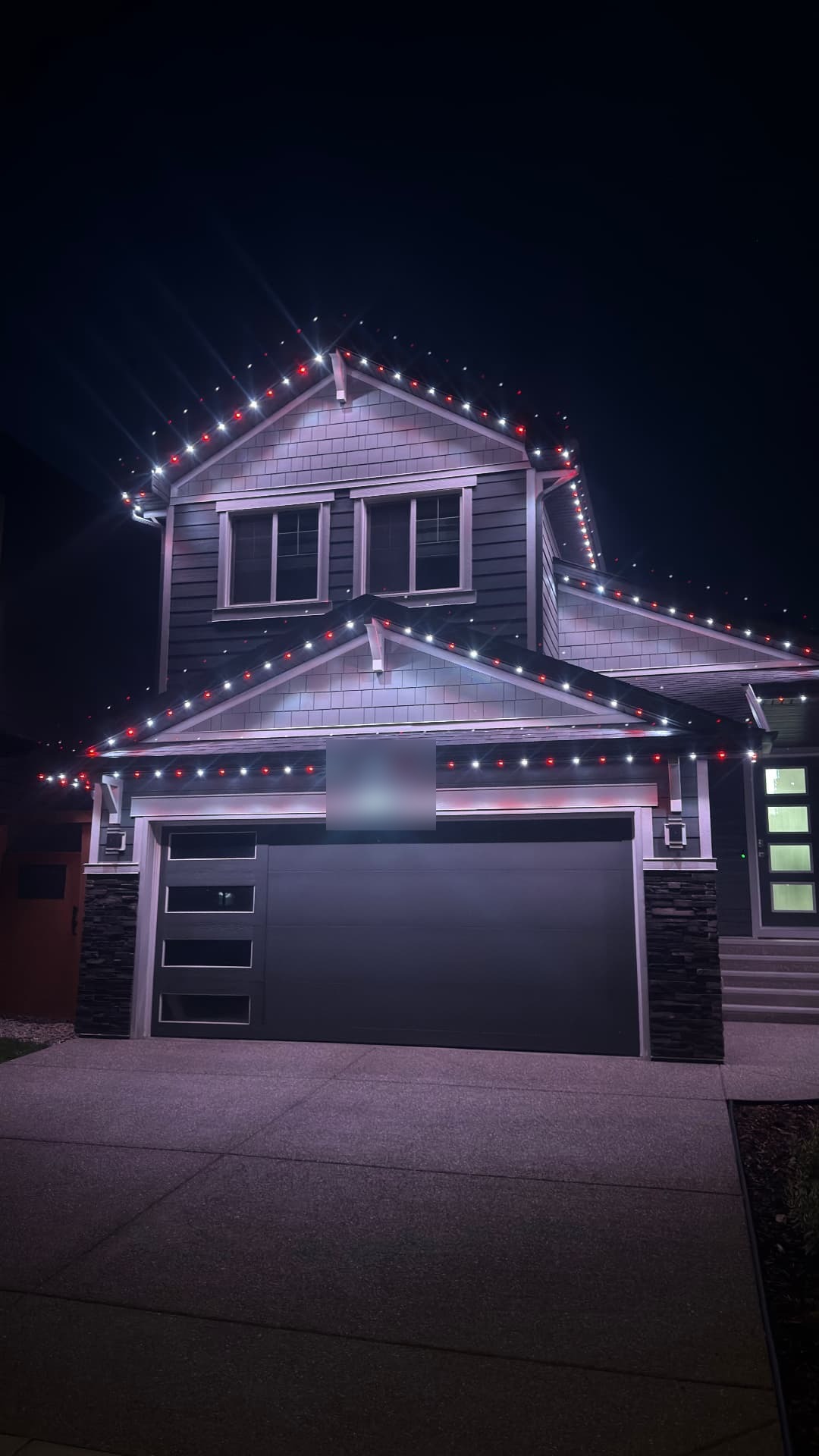 Two-storey home at night with alternating red and white permanent lights following every gable peak, eave and the garage line. House number digitally blurred for privacy.