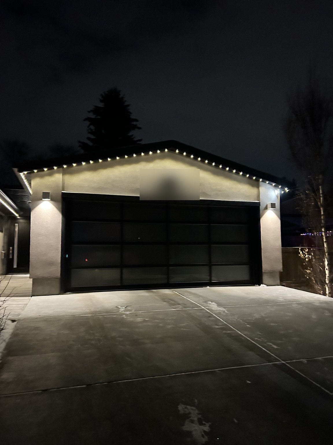 Detached garage at night with an even run of crisp warm white permanent lights following the full roofline above a dark glass garage door. House number digitally blurred for privacy.