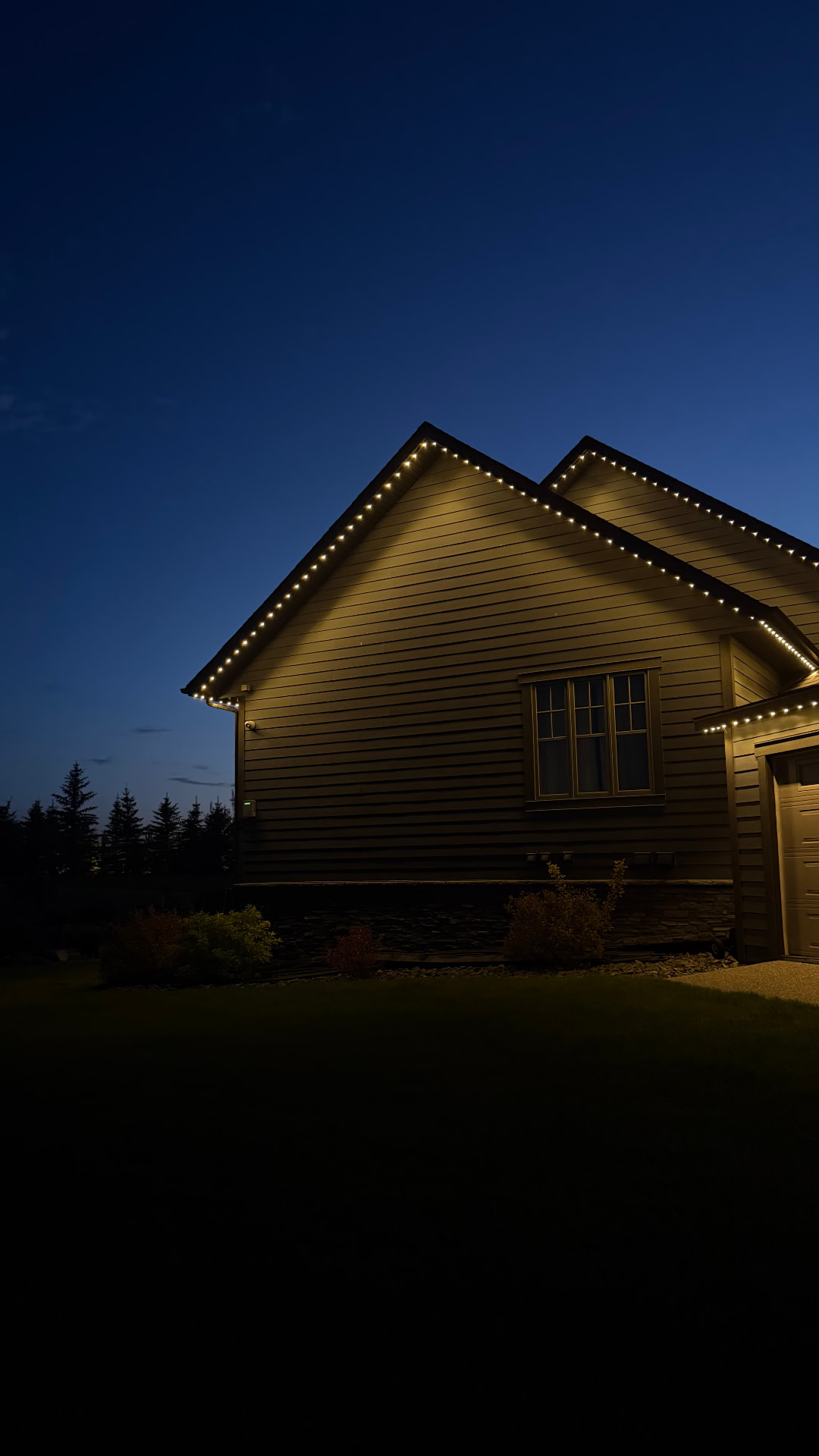Side elevation of a two-gable home photographed at deep twilight, with evenly spaced warm white permanent lights tracing both gable peaks and continuing along the eave to the garage.