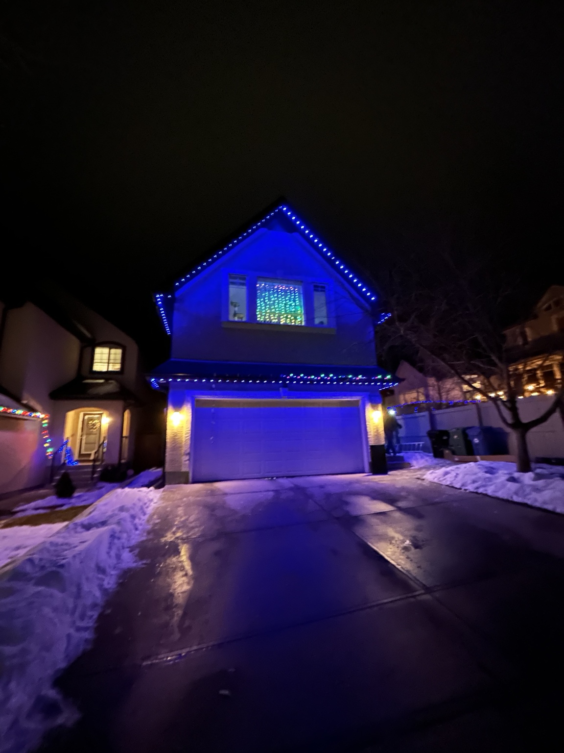 Two-storey Calgary home at night with permanent roofline lighting set to a deep blue scene along the peak, eaves and garage line, snow on the driveway.
