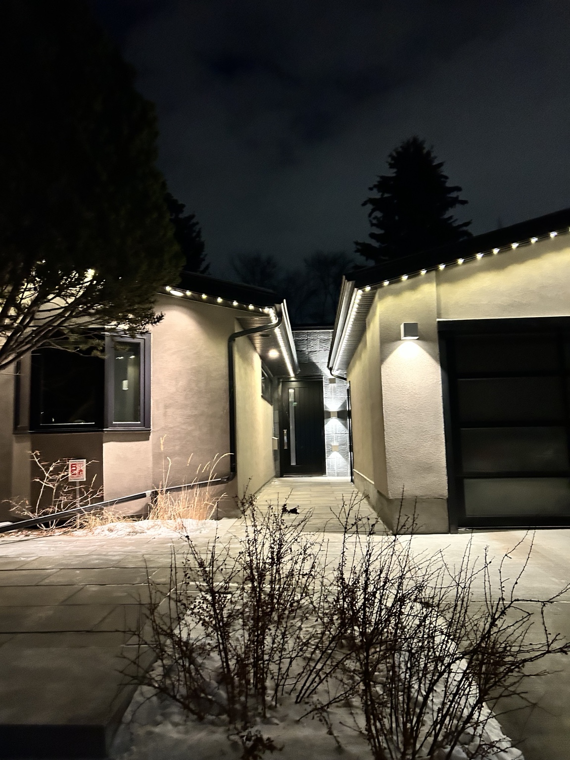 Modern bungalow entry courtyard at night, with warm white permanent lights following the low rooflines on both sides of a paved walkway leading to the front door.